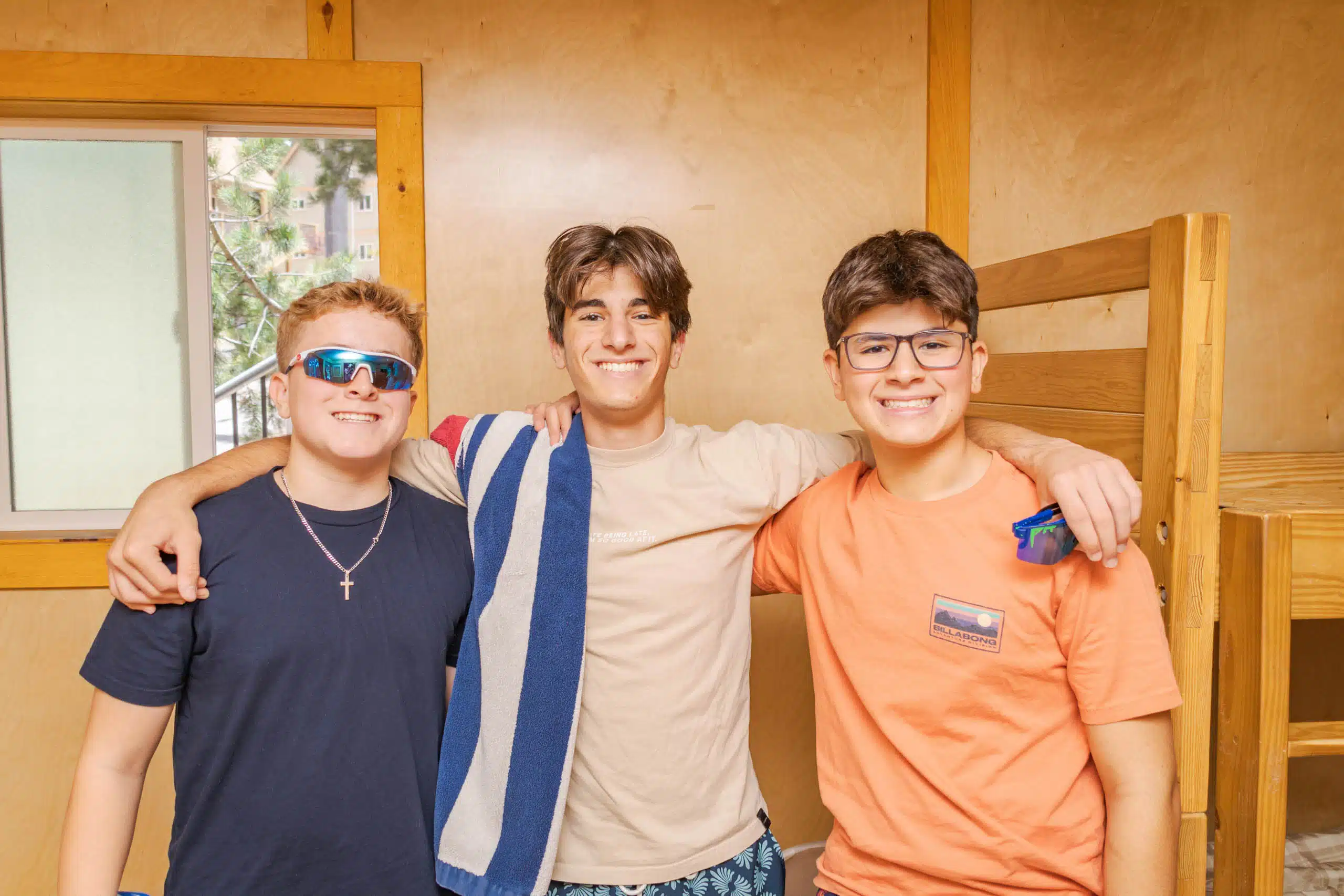 Three young men smiling, standing with arms around each other in a wooden room. Two wear glasses, one has a towel over his shoulder.