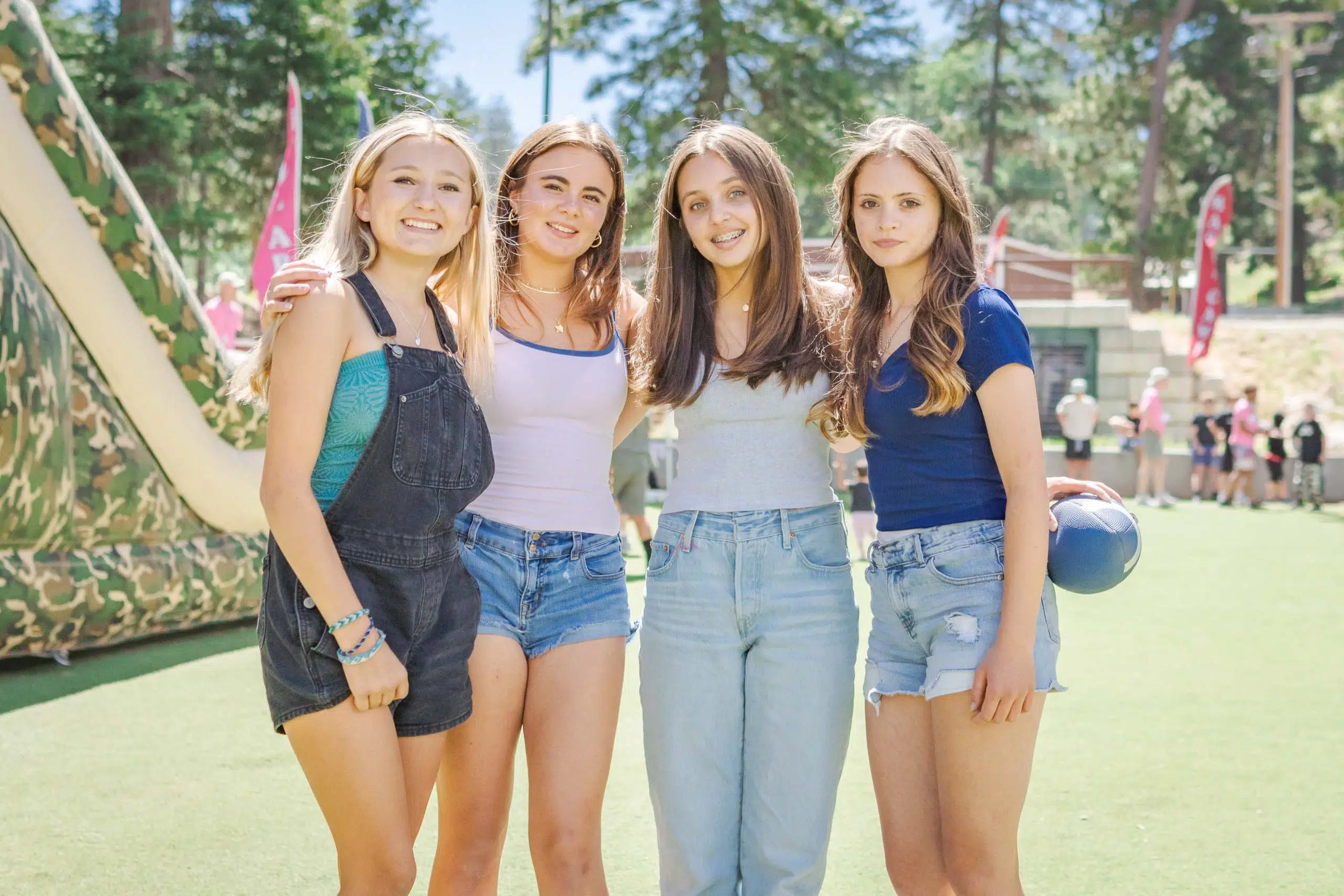 Four young women stand outdoors on grass in front of an inflatable slide, wearing casual summer clothes, with trees and people in the background.