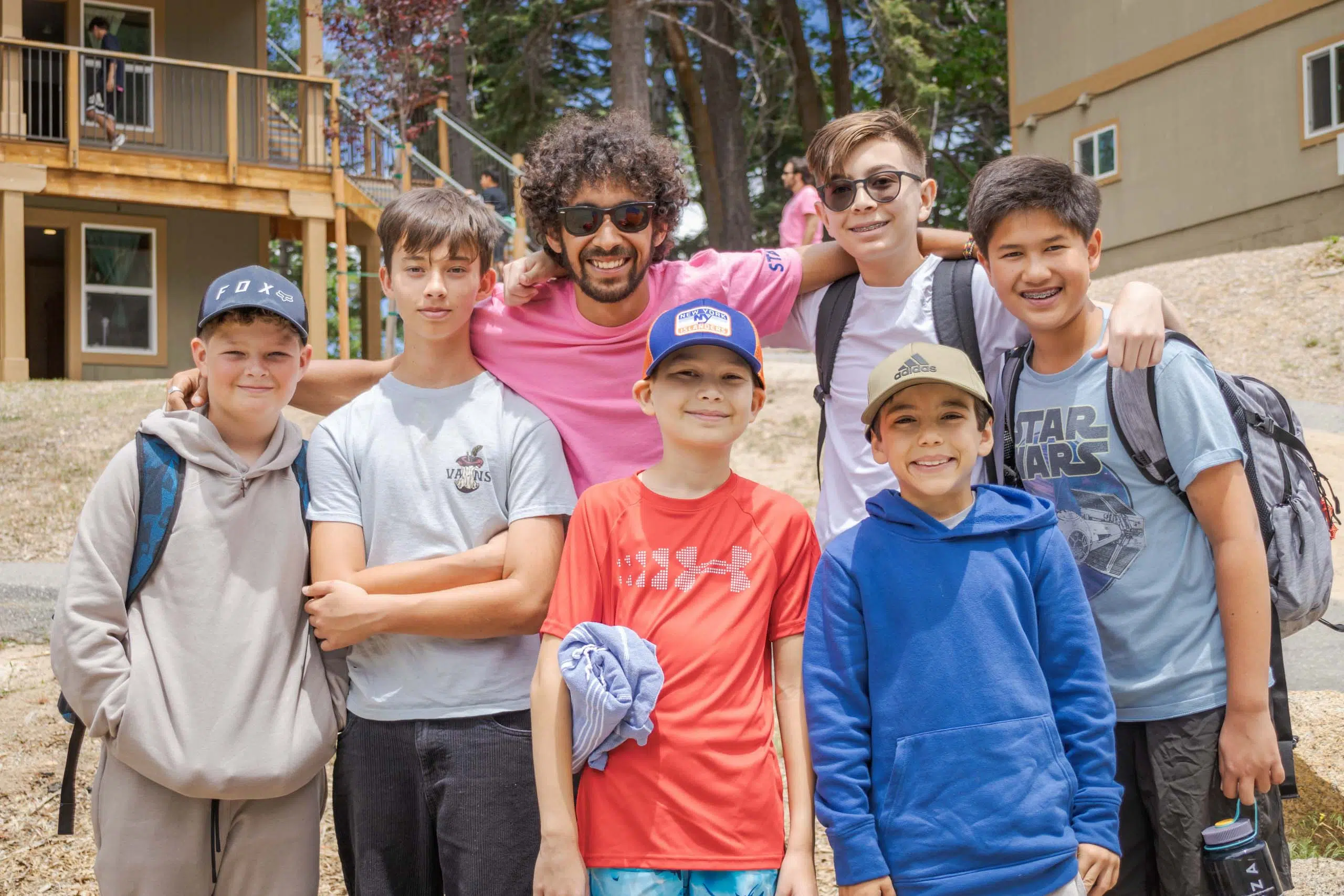 A group of seven people, including one adult and six children, pose outdoors with trees and buildings in the background.