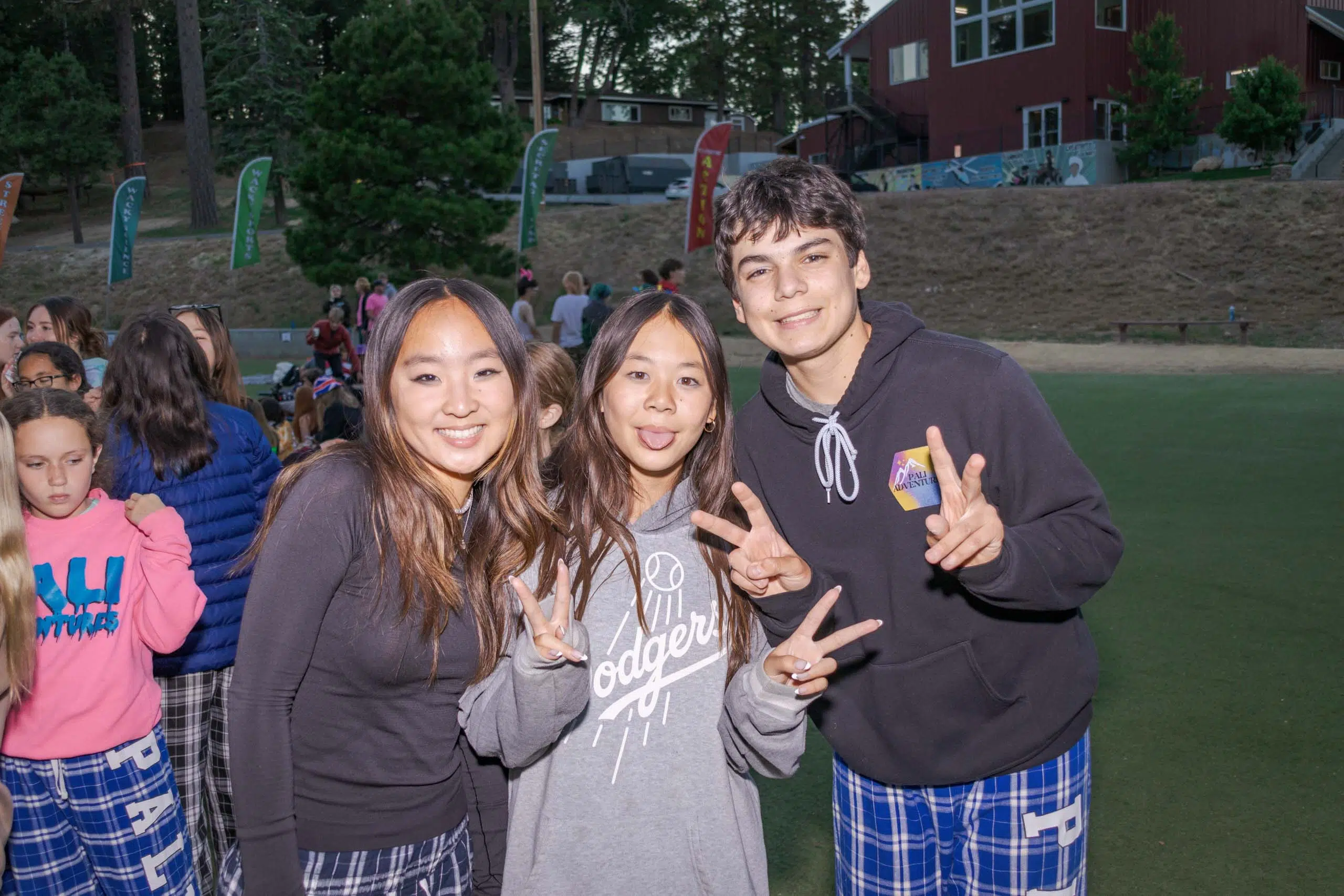 Three teenagers pose with peace signs on a grassy area in front of a building, surrounded by a group of people.