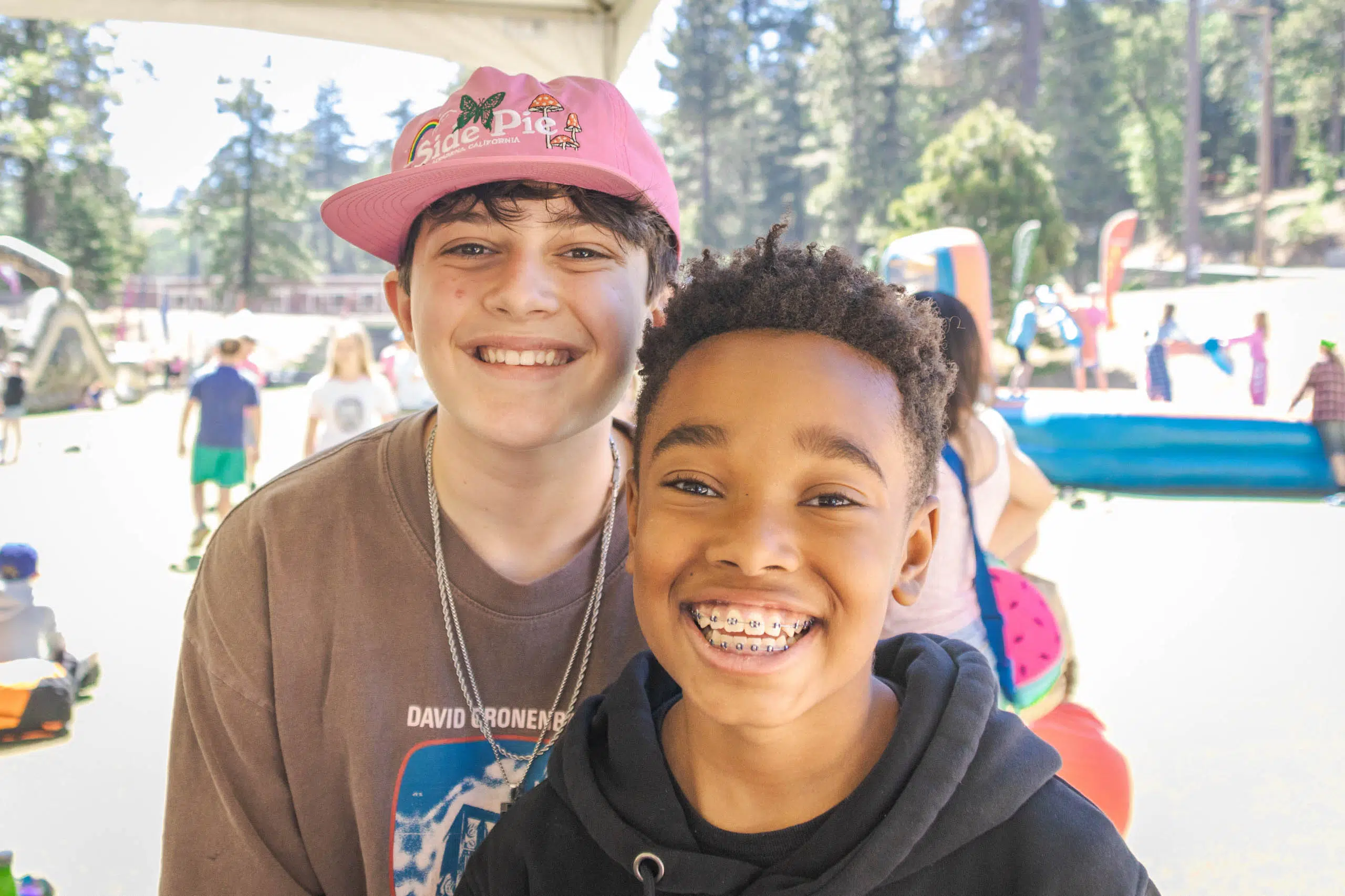 Two smiling boys stand outdoors under a tent. One wears a pink hat and brown t-shirt, the other has a black hoodie and braces. Behind them are trees and people at an event.