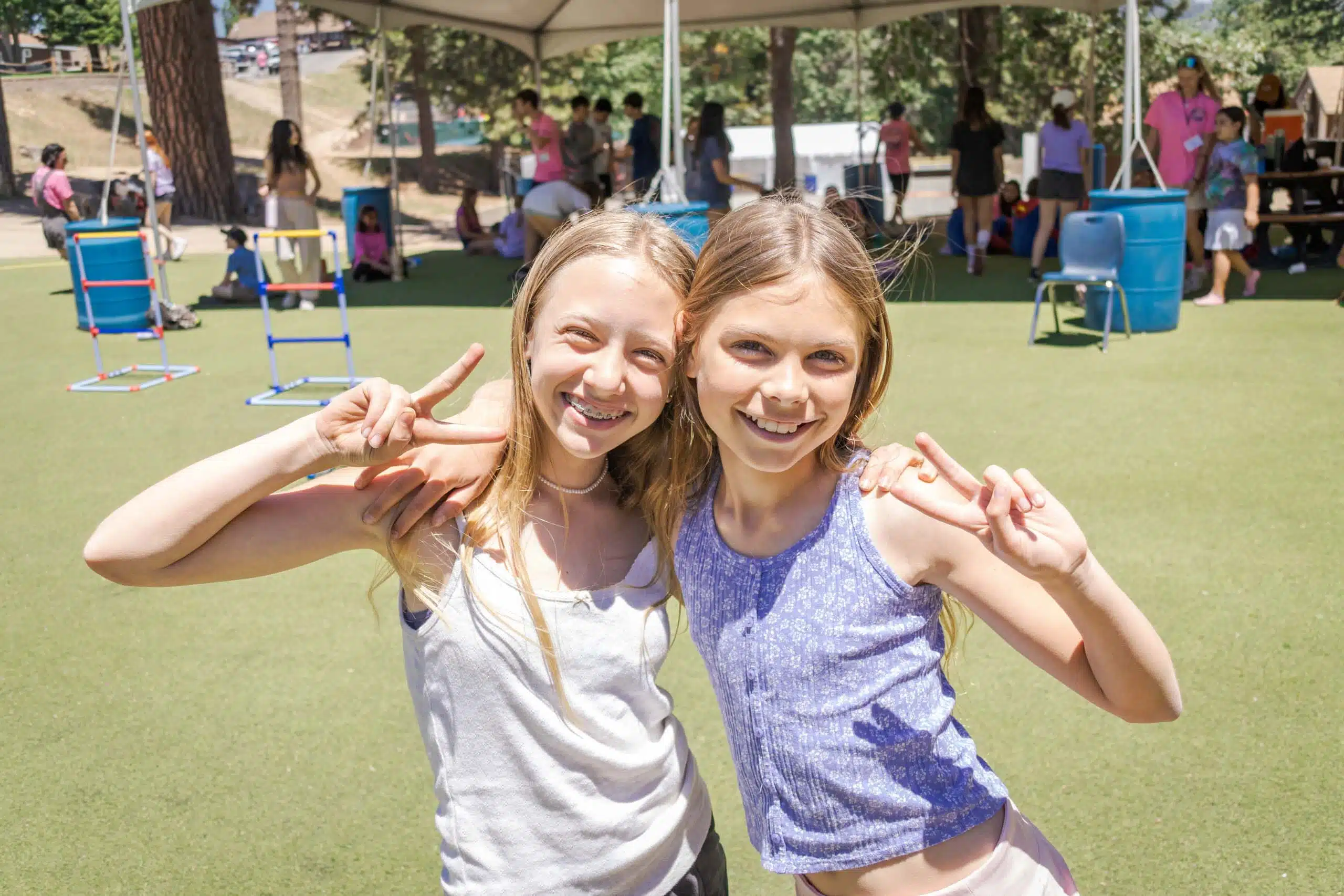 Two smiling children stand arm in arm on a grassy area under a canopy, with people and outdoor equipment in the background.